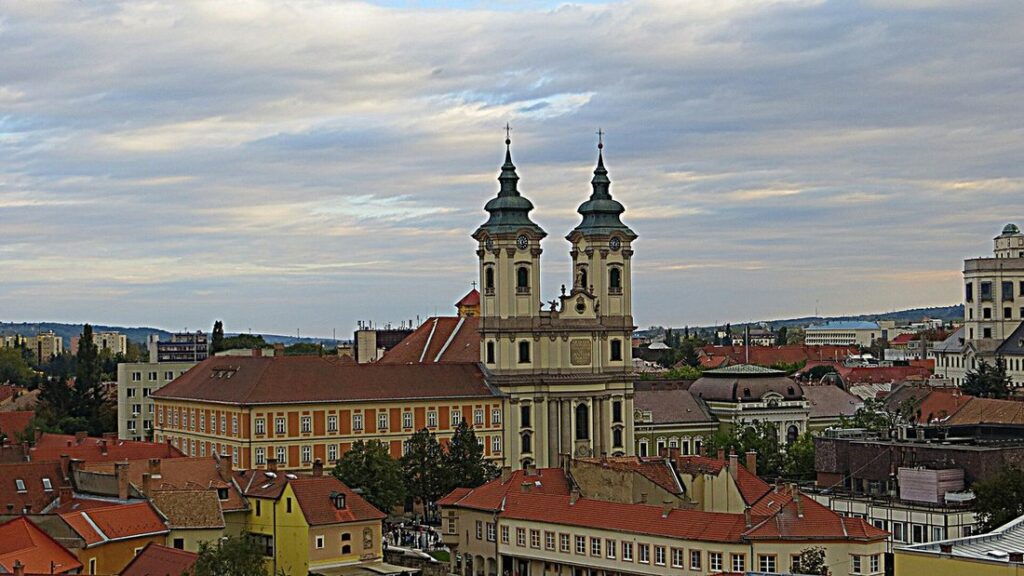 Clochers jumeaux d'une église baroque dominant les toits de tuiles rouges d'une ville, sous un ciel nuageux