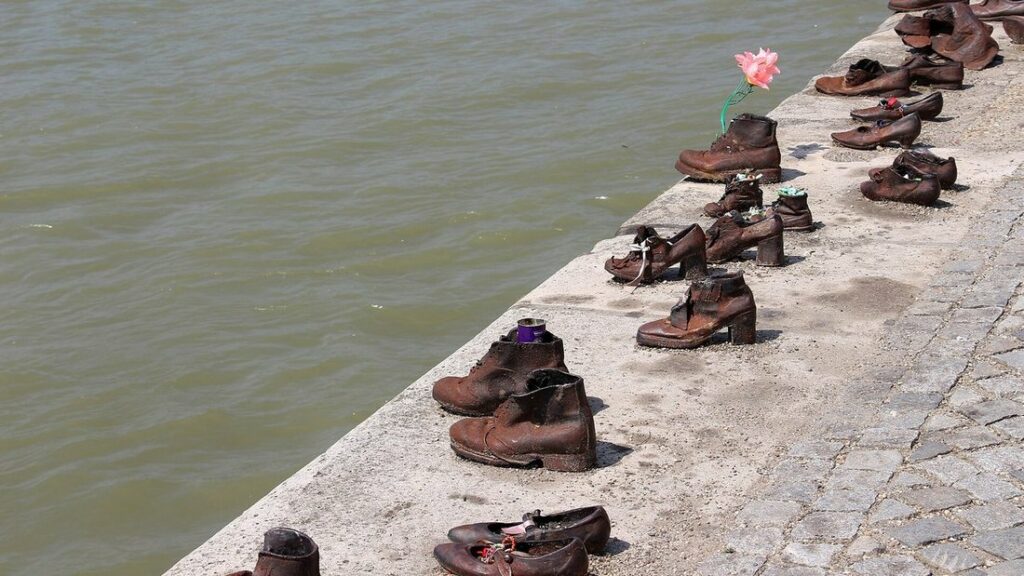 Rangée de chaussures en bronze alignées sur le quai de pierre, avec le Danube derrière et une fleur rose posée dessus