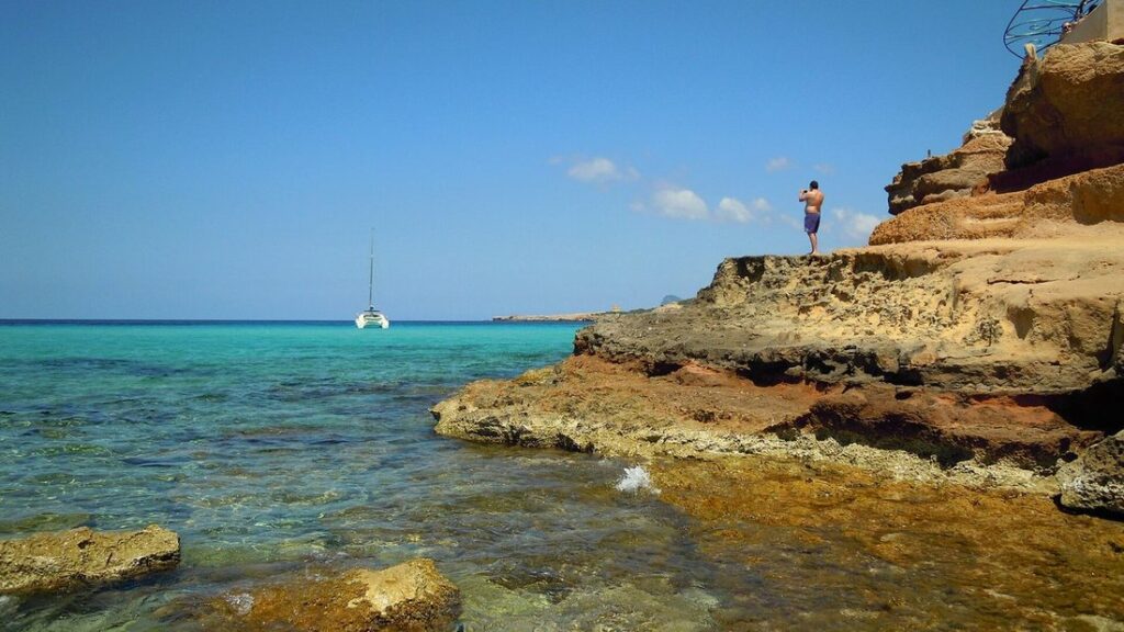 Eau turquoise limpide au pied de rochers ocre à Cala Comte, avec un voilier au large