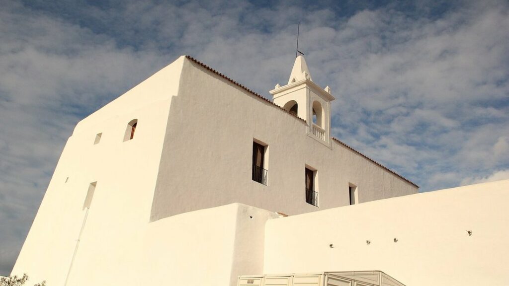 Murs blancs chaulés et petit clocher de l'église de Sant Miquel de Balansat, vus d'en bas sous des nuages légers