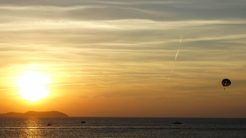 Soleil bas et orangé sur la mer, avec un îlot en silhouette à l'horizon et un parachute dans le ciel à droite