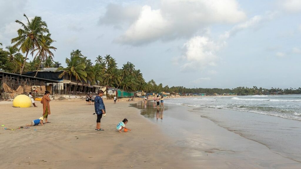 Plage de sable clair bordée de cocotiers, avec quelques promeneurs et la mer à droite