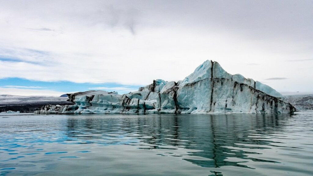 Iceberg rayé de noir et de bleu qui flotte sur l'eau calme d'une lagune, sous un ciel dégagé