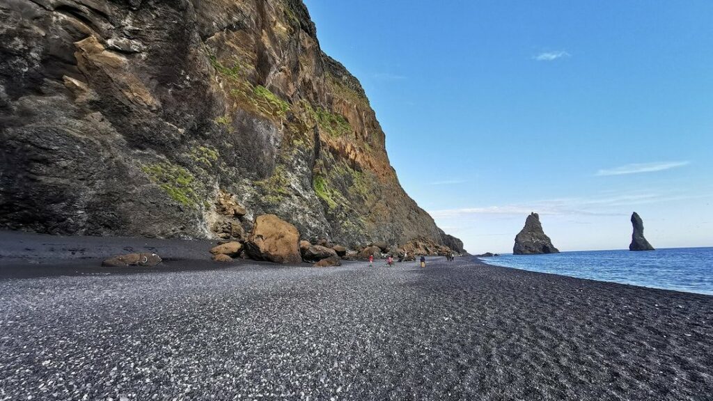 Plage de galets et de sable noir au pied d'une falaise brune, avec deux aiguilles rocheuses au bord de la mer
