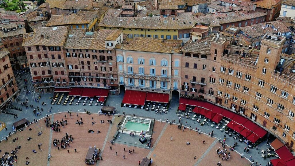 Vue plongeante sur une grande place pavée bordée de palais de brique, avec des terrasses à auvents rouges