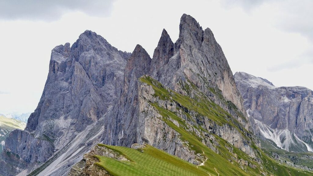 Crêtes rocheuses déchiquetées dominant des prairies vertes en pente, sous un ciel voilé