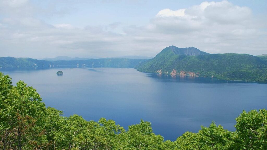 Grand lac bleu entouré de pentes boisées vertes, avec un relief volcanique au fond sous un ciel nuageux