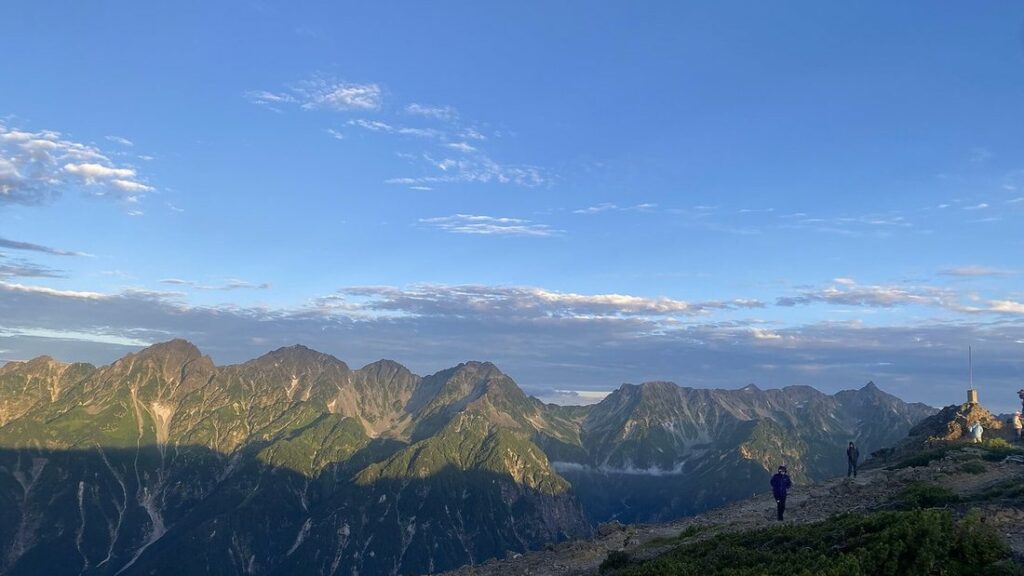 Crêtes rocheuses et sommets des Alpes japonaises, avec deux randonneurs sur un sentier sous un ciel bleu