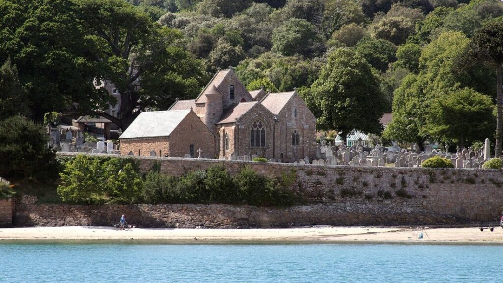Église en pierre au bord d'une plage de sable, entourée d'arbres, avec une eau turquoise au premier plan