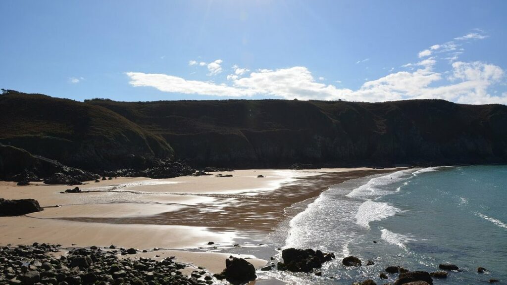 Plage de sable ourlée d'écume au pied de hautes falaises sombres, sous un ciel bleu nuageux