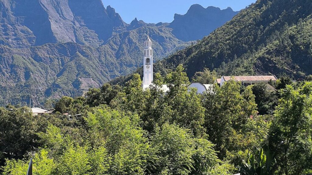 Clocher blanc d'une église parmi les arbres, au pied de pitons rocheux et de pentes vertes