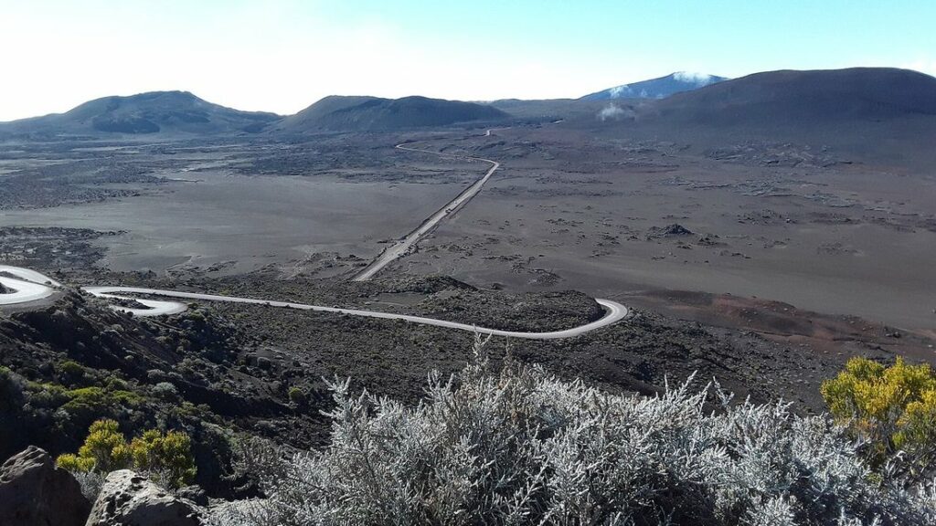 Route sinueuse traversant une vaste plaine de lave grise, avec des reliefs au loin sous un ciel bleu