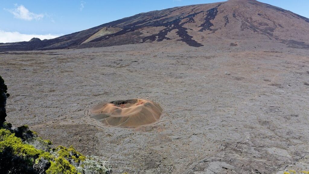 Cône volcanique sombre dominant un champ de lave refroidie, avec un petit cratère orangé au premier plan
