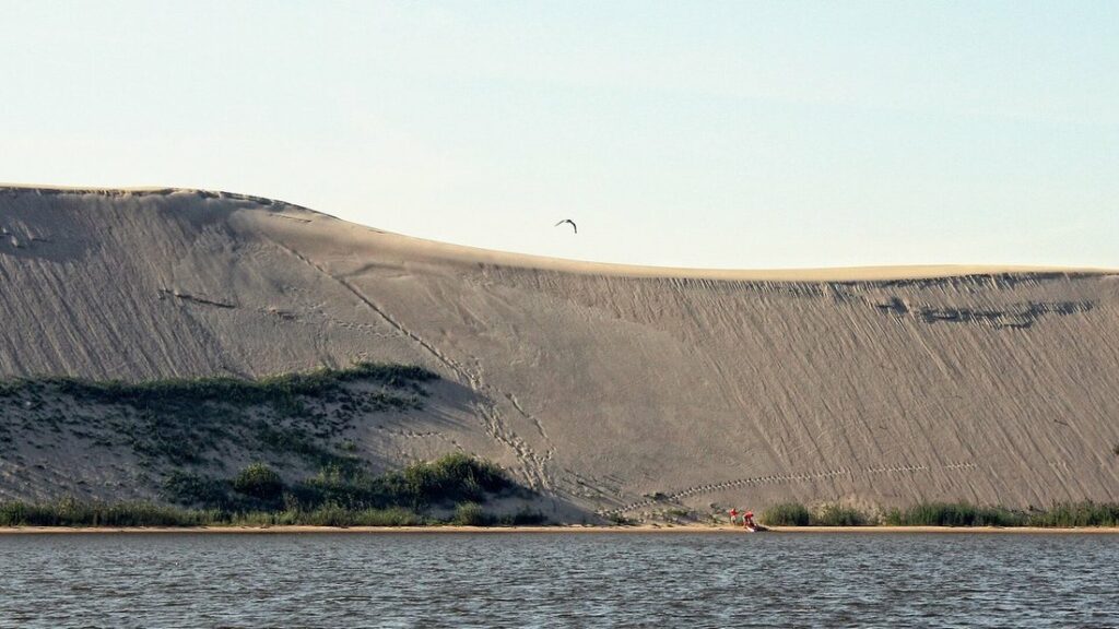 Grande dune de sable nu descendant vers l'eau du lagon, sous un ciel clair