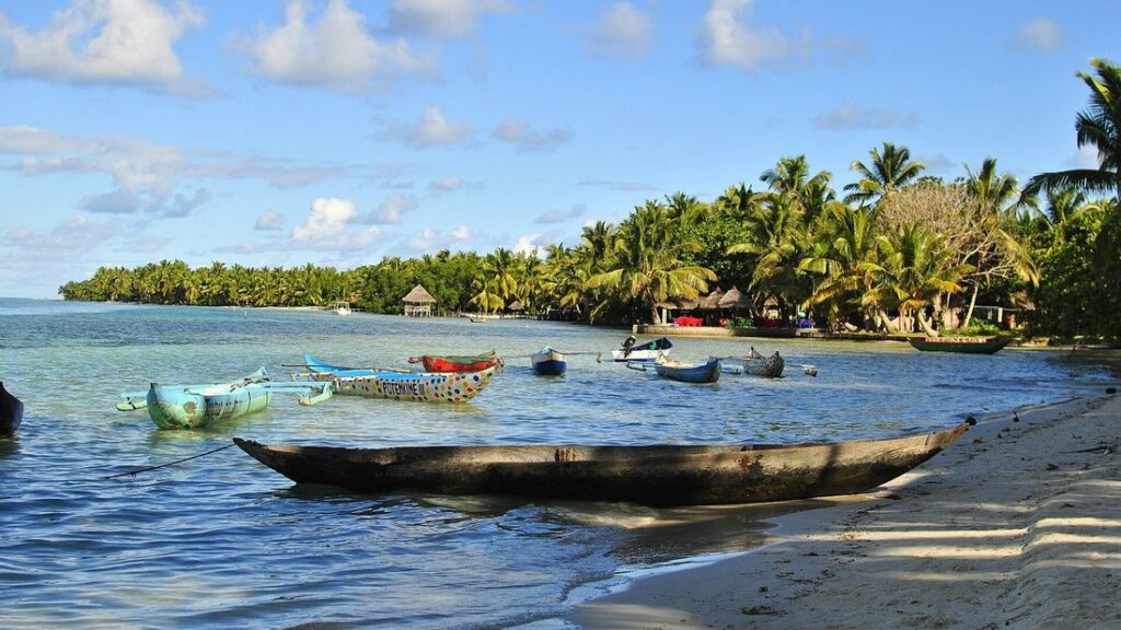 Pirogues en bois sur le sable et dans l'eau d'une baie bordée de cocotiers, sous un ciel bleu nuageux