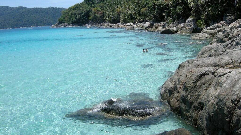 Eau turquoise limpide au pied de rochers arrondis, avec une côte boisée en arrière-plan