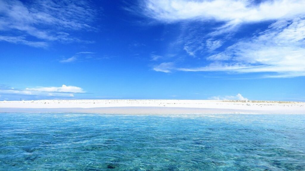 Longue bande de sable blanc à fleur d'eau au milieu d'un lagon turquoise, sous un ciel bleu et nuageux