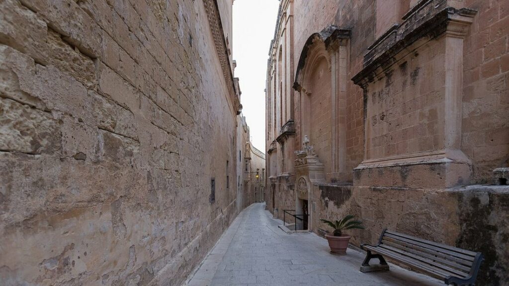 Ruelle étroite de Mdina entre de hauts murs de pierre claire, avec un banc et une pente pavée