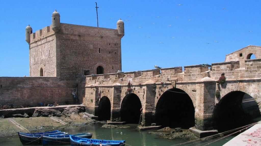 Bastion et remparts de pierre aux arches côté port d'Essaouira, avec des barques bleues au pied et un ciel bleu