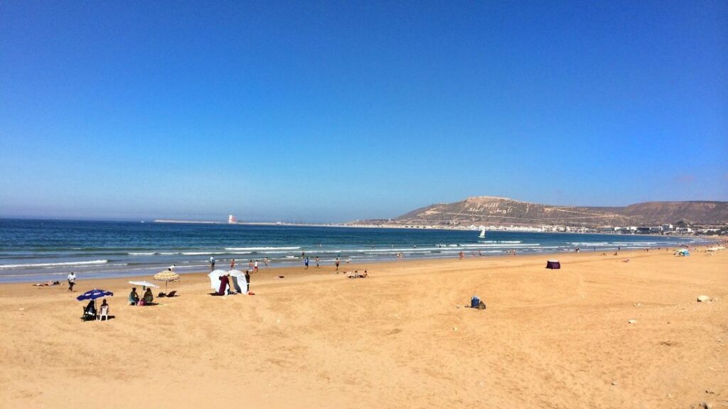 Plage de sable doré avec quelques parasols, mer bleue et colline dominant la baie sous un ciel bleu