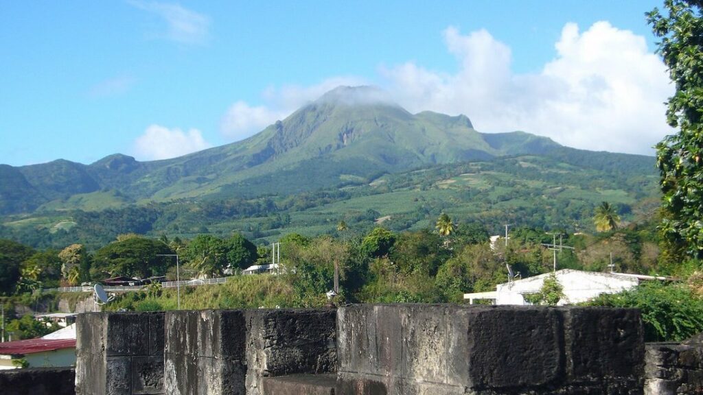 Montagne verte au sommet voilé de nuages, au-dessus de collines cultivées, avec un mur de pierre sombre au premier plan