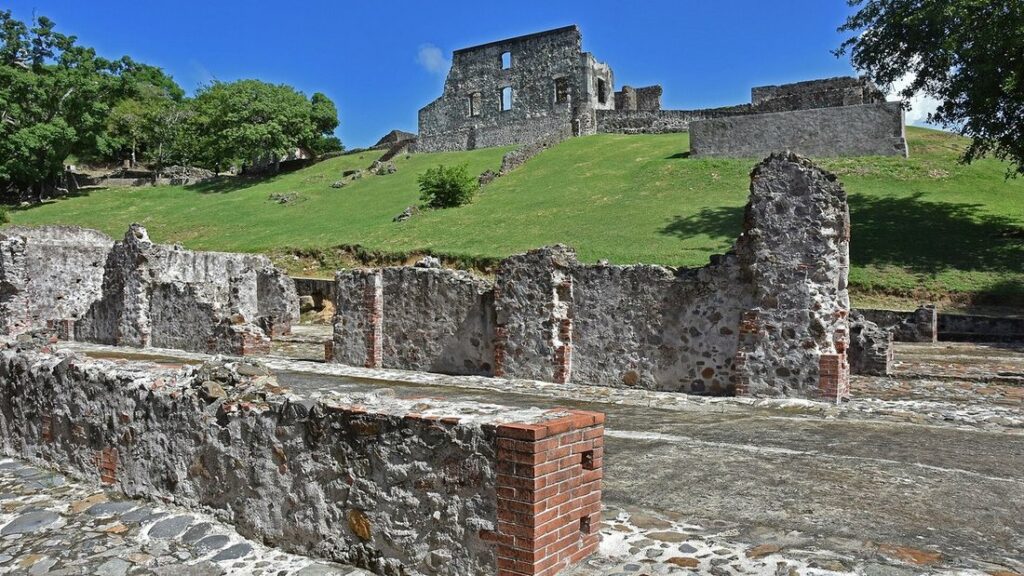 Murs de pierre en ruine sur une pelouse verte, avec un grand pan de mur resté debout au sommet de la butte