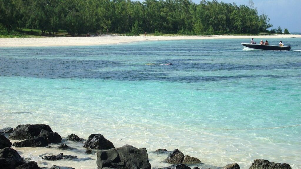 Eau turquoise peu profonde, rochers noirs, plage de sable clair et bateau rapide devant une rangée d'arbres