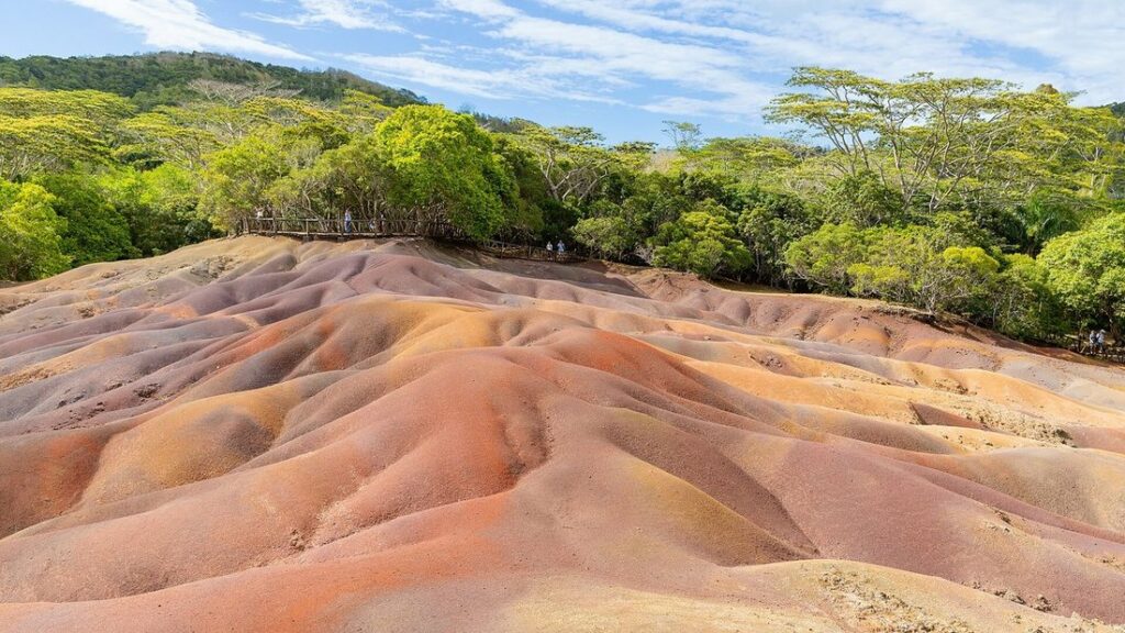 Dunes de terre aux teintes roses, orangées et beiges, bordées d'arbres sous un ciel partiellement nuageux