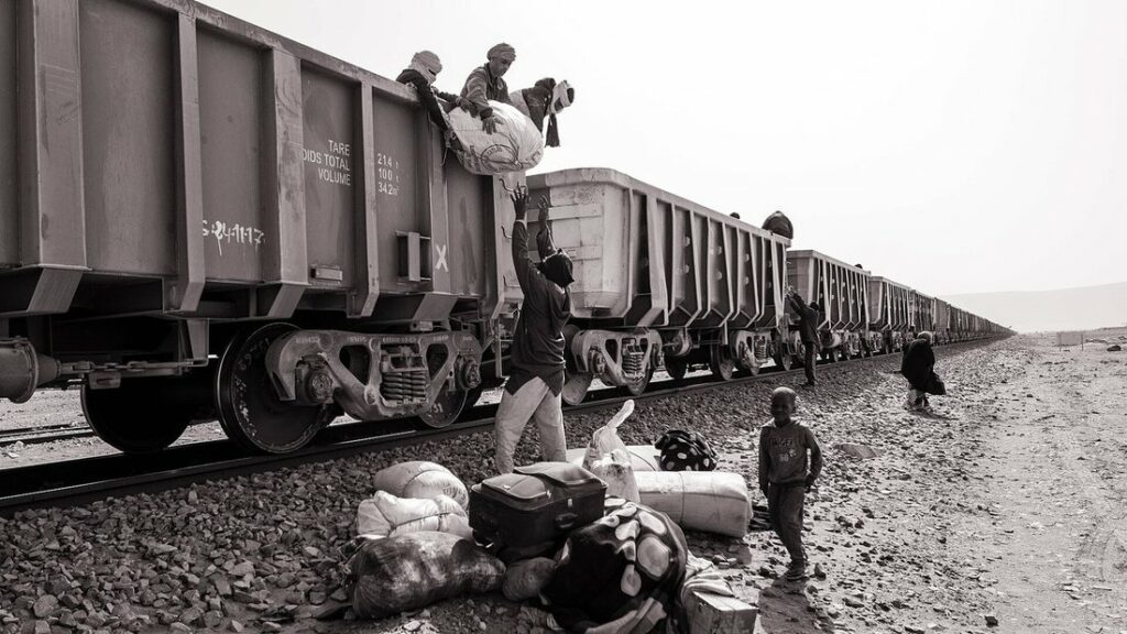 Train de wagons à minerai sur une voie dans le désert, avec des voyageurs et leurs bagages au bord