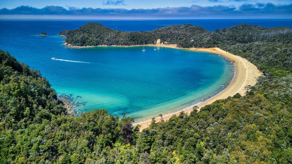 Vue aérienne d'une baie en croissant, avec une plage de sable clair, une eau turquoise et des collines boisées