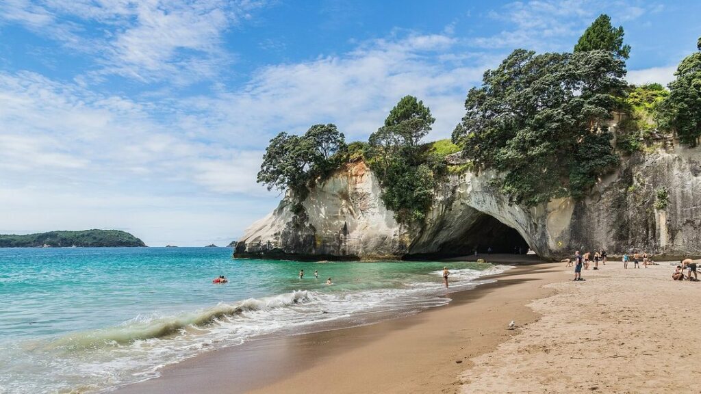 Arche de falaise blanche couronnée d'arbres au bord d'une plage de sable, avec des vagues et quelques promeneurs