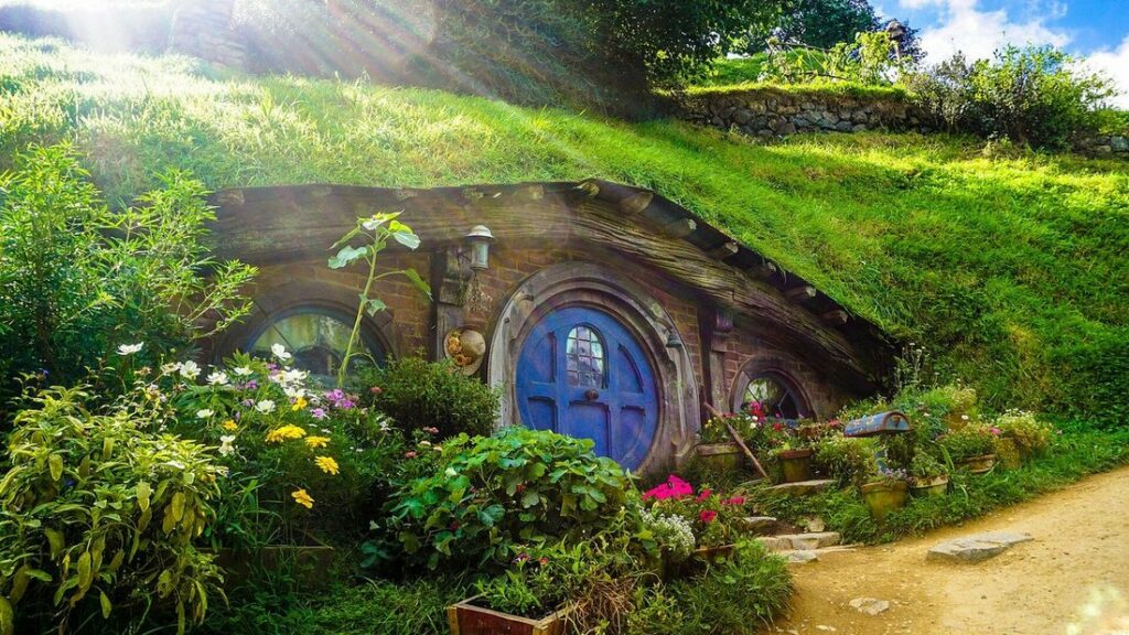 Maison de hobbit semi-enterrée à porte ronde bleue, entourée de fleurs et d'herbe verte