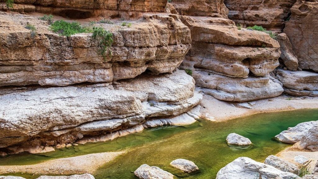 Strates de roche claire et vasque d'eau verte entre des blocs lisses dans le Wadi Shab