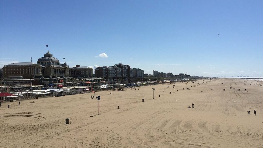 Large plage de sable presque vide sous un ciel bleu, avec le Kurhaus et la promenade au fond