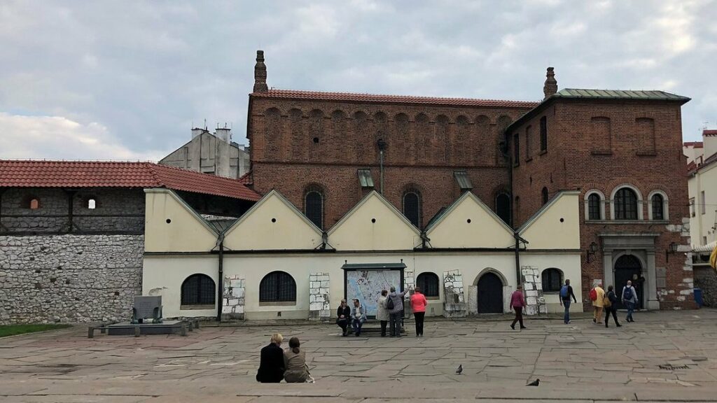 Façade de brique d'une ancienne synagogue, précédée de bâtiments bas à pignons blancs, sur une place pavée