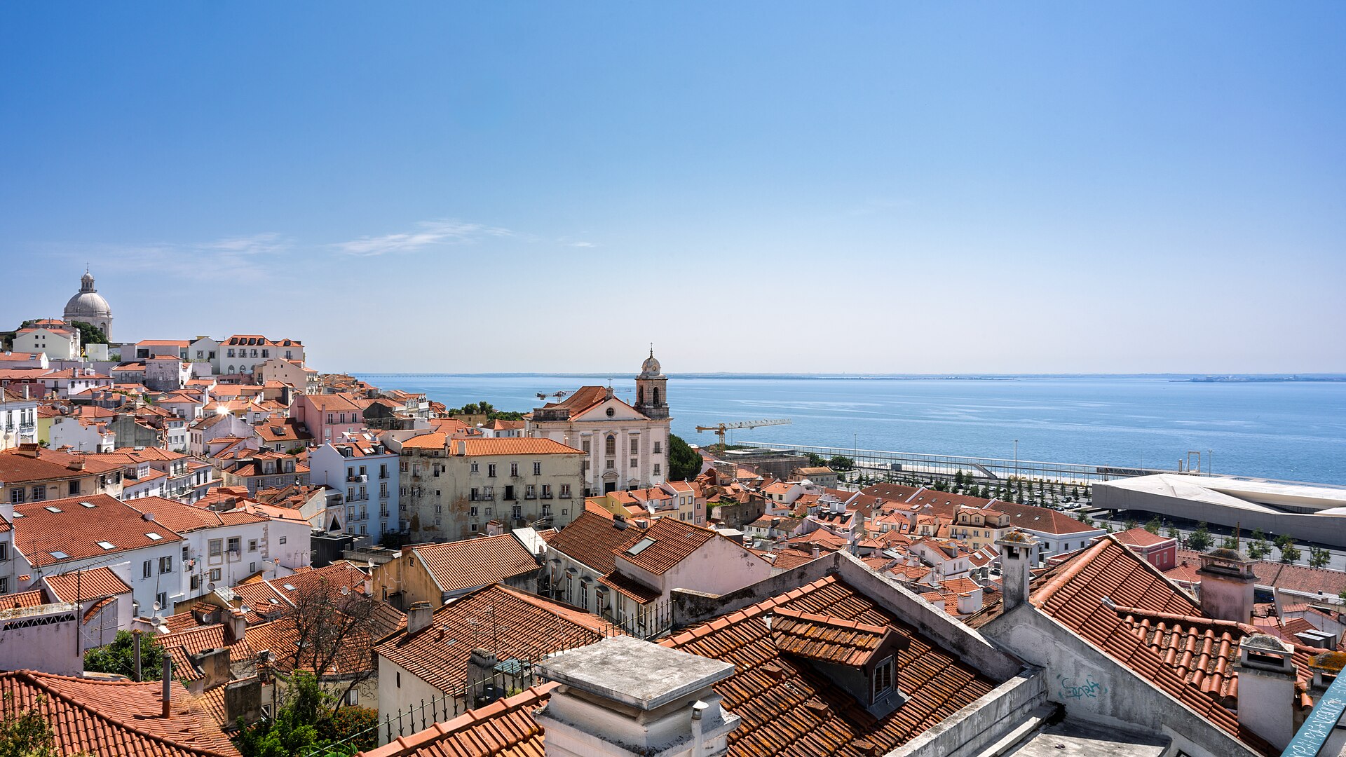 Toits de tuiles rouges de l'Alfama, clocher d'église et dôme blanc, avec l'estuaire du Tage sous un ciel clair