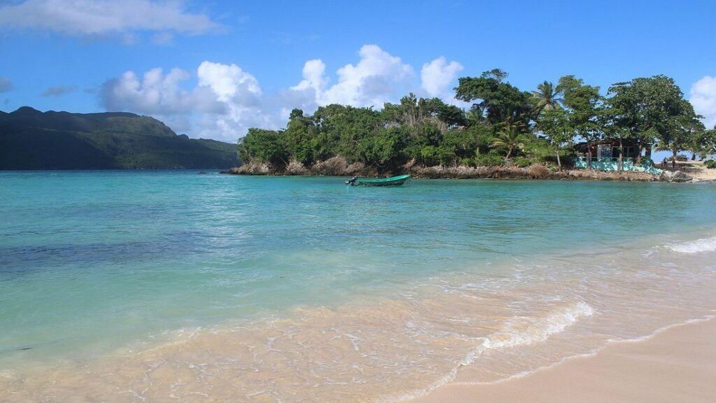Plage de sable clair, eau turquoise et îlot boisé devant des collines vertes sous un ciel bleu