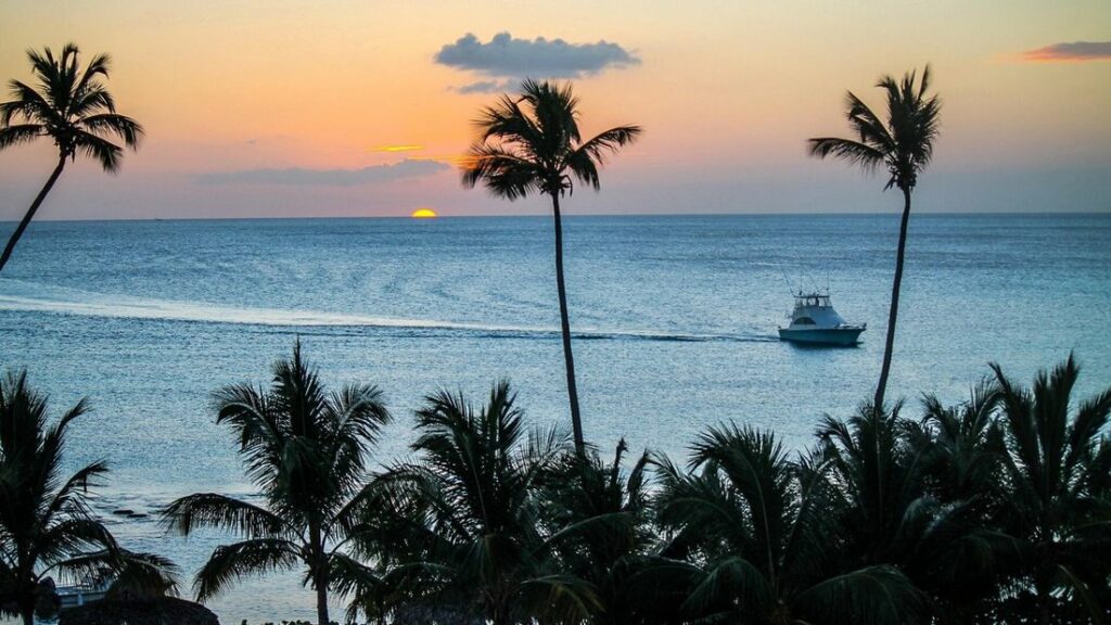 Palmiers découpés devant une mer calme où file un bateau, sous un ciel orangé près de l'horizon