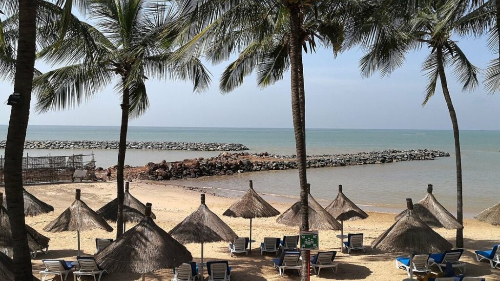 Plage de sable bordée de cocotiers, avec des parasols de paille et une digue de pierres dans la mer