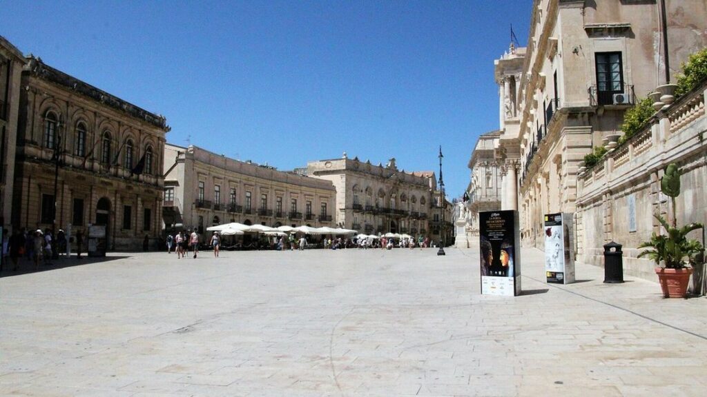 Grande place pavée d'Ortigia, bordée de façades claires et de terrasses, sous un ciel bleu