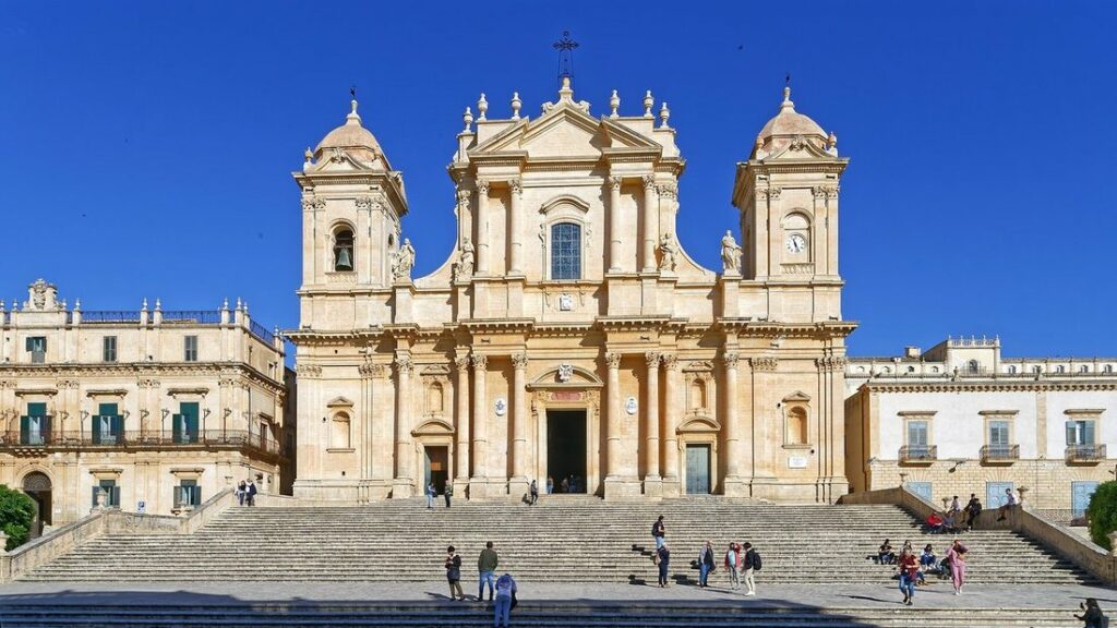 Façade baroque en pierre dorée de la cathédrale de Noto, au sommet d'un grand escalier, sous un ciel bleu