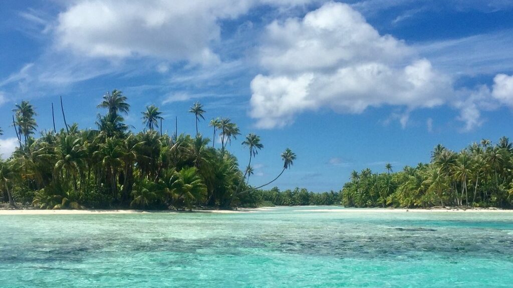 Lagon turquoise bordé d'un îlot de cocotiers, sous un ciel bleu et de gros nuages