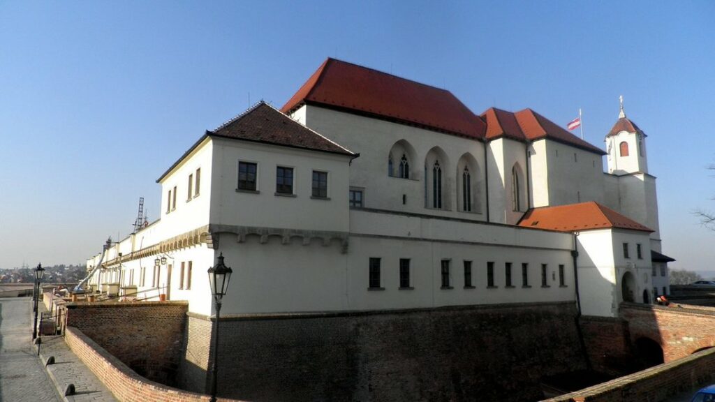 Château du Spielberk à Brno, grand bâtiment blanc à toit rouge dominant un rempart de brique, sous un ciel bleu