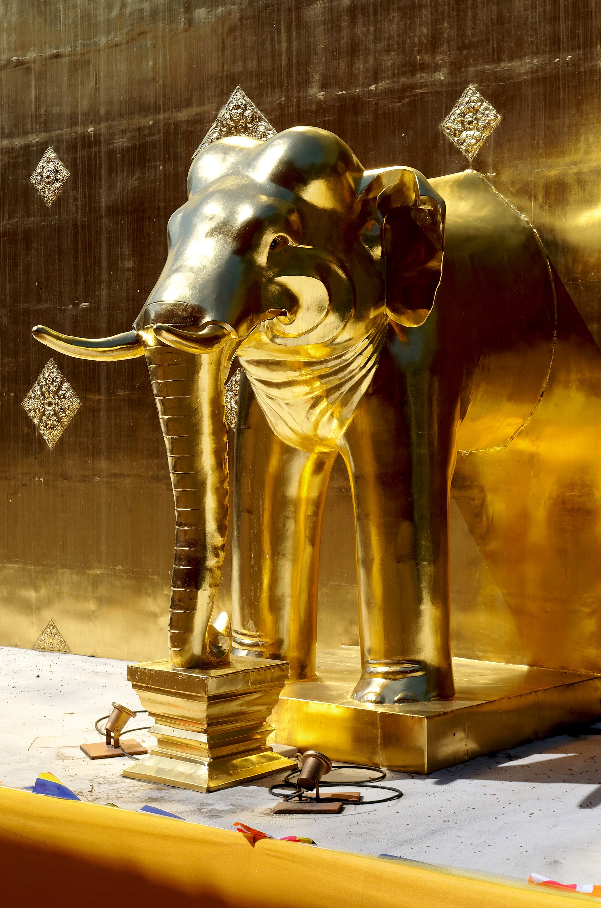 Statue d'éléphant dorée, trompe posée sur un socle, devant la paroi dorée d'un monument ornée de losanges