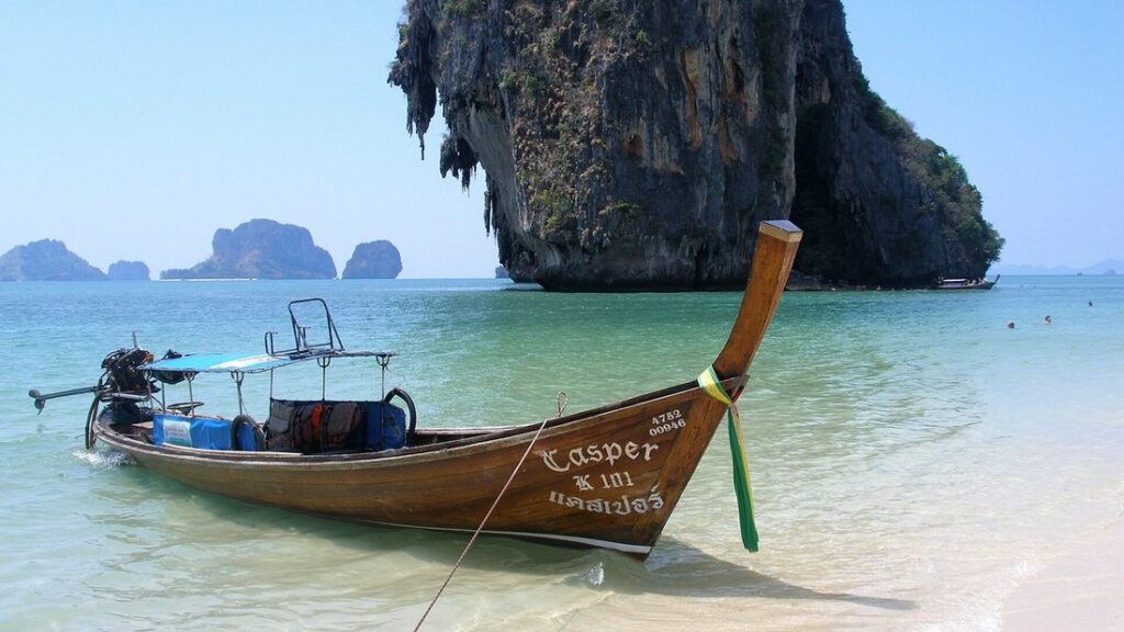 Bateau longtail en bois dans l'eau claire d'une plage de sable, devant une haute falaise calcaire