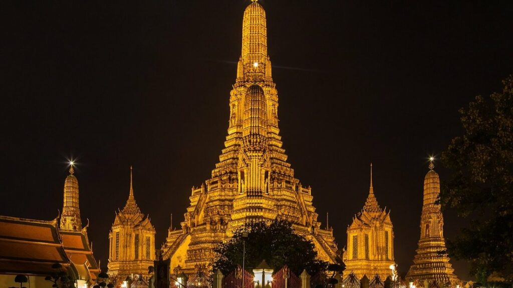 Haute tour dorée du Wat Arun éclairée de nuit, entourée de tours plus petites, sur un ciel noir
