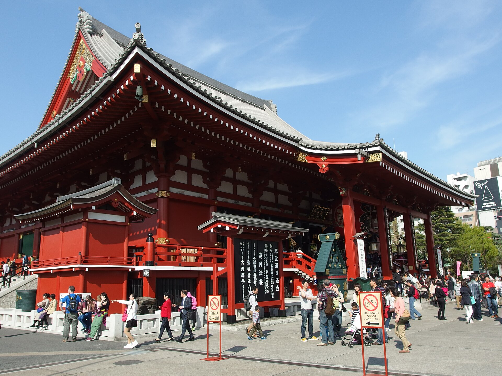 Grande salle principale rouge au toit de tuiles grises, avec des visiteurs sur l'esplanade sous un ciel bleu