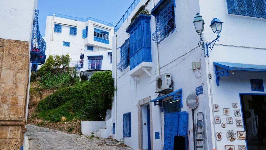Façades blanches aux volets et balcons bleus le long d'une ruelle en pente, avec un réverbère et de la verdure