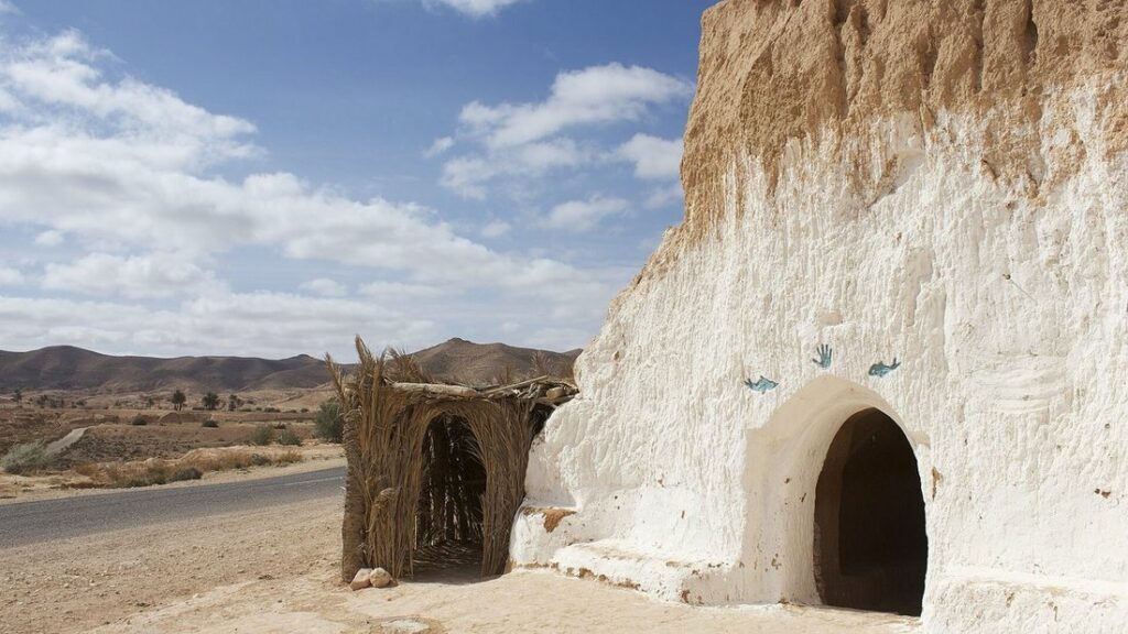 Habitation creusée dans une falaise de roche claire, avec une porte voûtée, une cabane de palmes et une route