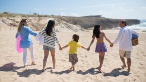 Une famille de cinq personnes vue de dos, main dans la main, qui marche sur une plage de sable avec une bouée rose et un panier